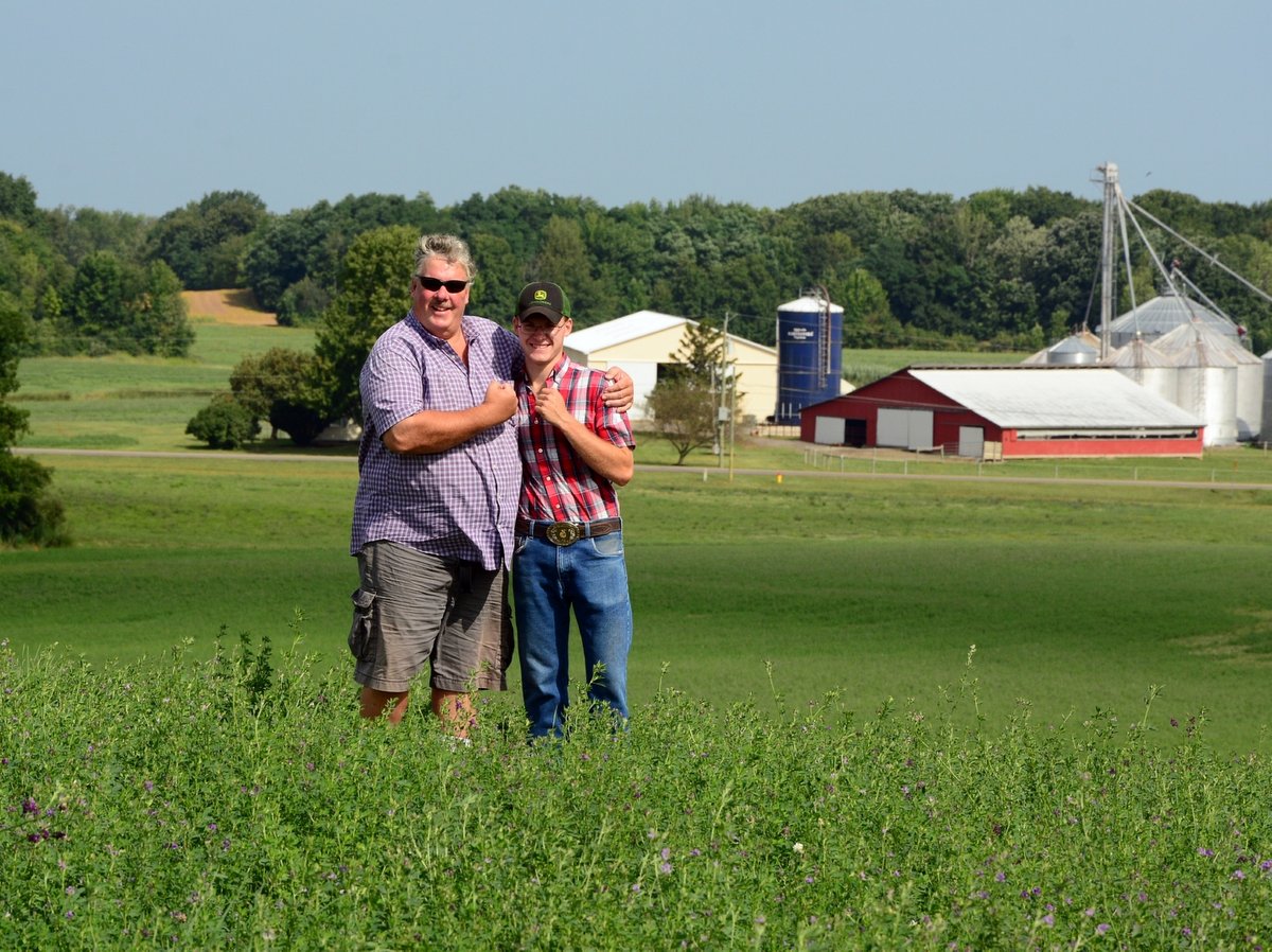 A father and son standing in a field with farm buildings in background