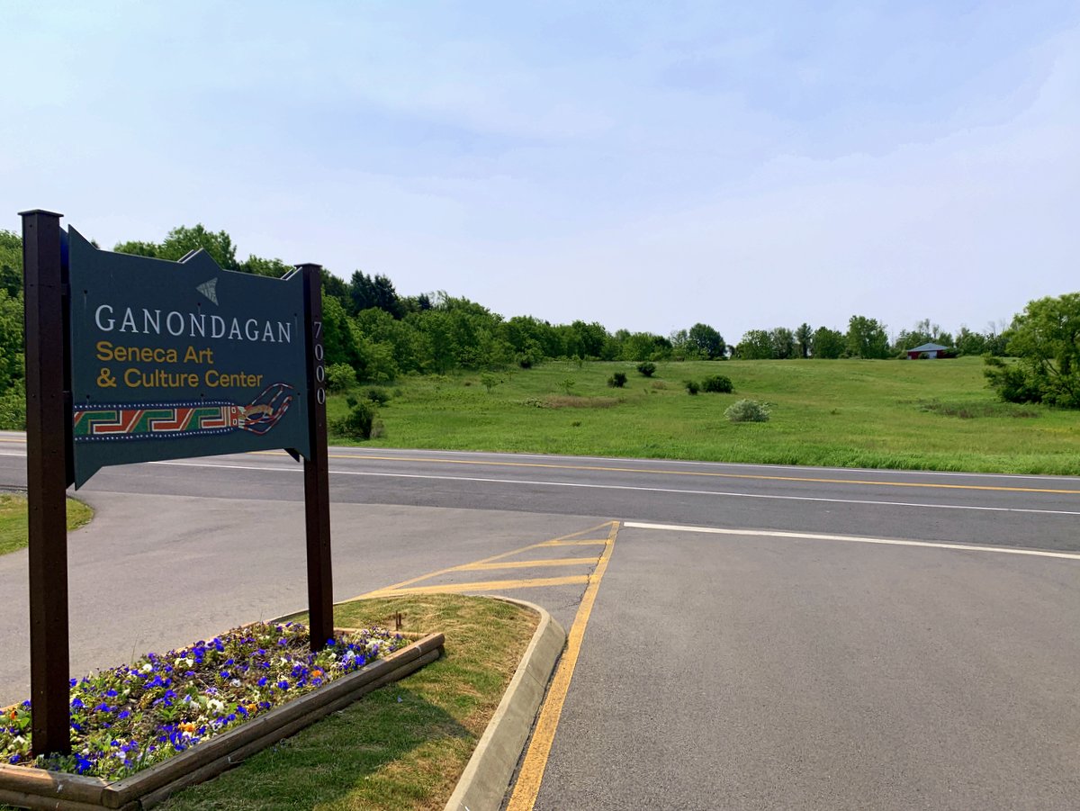 A green meadow across from the entrance to a parking lot with a sign indicating "Ganondagan State Historic Site"